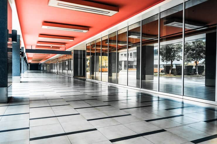 Modern office with floor-to-ceiling glass partitions offering a panoramic view of a city skyline. The space features polished concrete flooring, an industrial-style ceiling with exposed ductwork, and pendant lighting. Natural light floods the room, creating a bright and open atmosphere