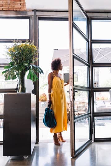 A stylish woman in a bright yellow dress holding a blue handbag stands at the open glass partition door of a modern entrance. The scene includes a black cabinet with a vase of fresh flowers and large outdoor umbrellas visible through the glass, creating an inviting and contemporary atmosphere