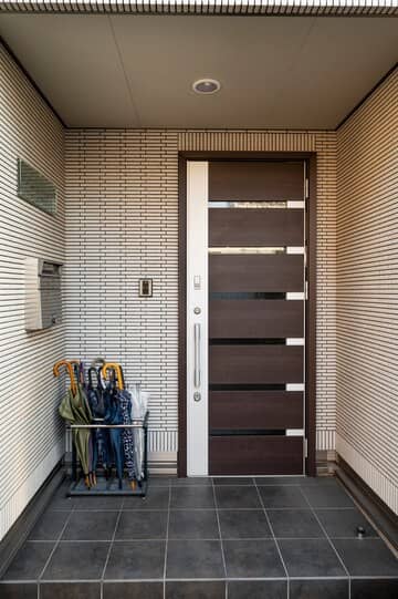 Contemporary entrance featuring a sleek wooden door with horizontal glass panels, set against a textured tiled wall. The area includes a black tiled floor and an umbrella stand with multiple umbrellas, adding a practical touch to the stylish design