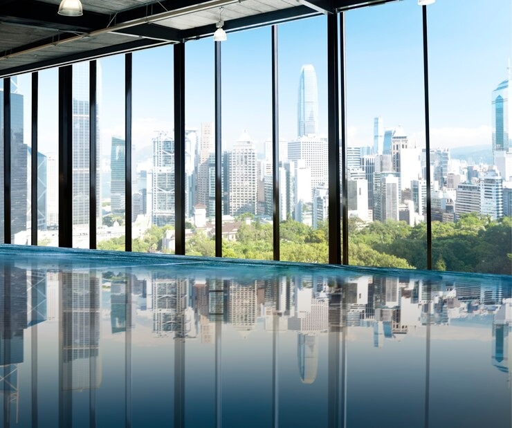 Modern office with floor-to-ceiling glass wall partitions offering a panoramic view of a city skyline and green park. The space features polished concrete flooring, industrial-style ceiling with exposed ductwork, and pendant lighting. Natural light floods the room, creating a bright and open atmosphere