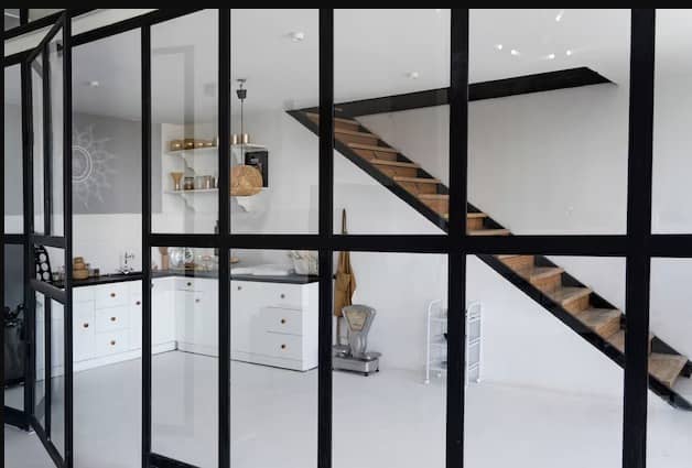 Contemporary kitchen area featuring sleek white cabinets, a floating wooden staircase, and glass partition walls with black frames. The space is accentuated with open shelving, gold accents, and minimalistic decor